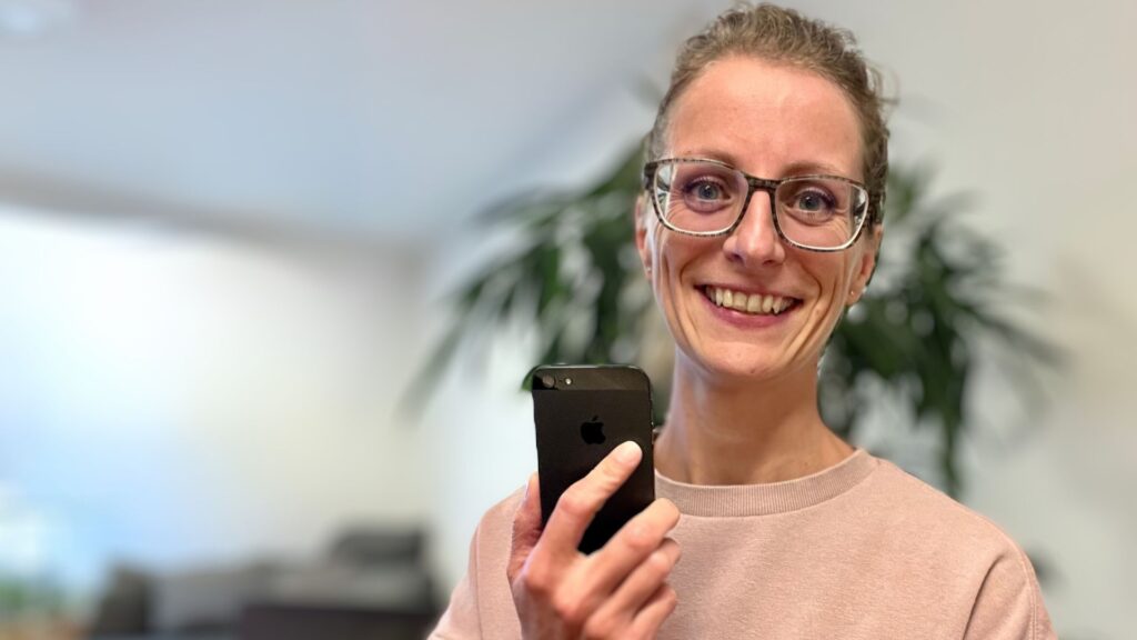 Instagram-Beraterin Martina Lörracher-Leeb lächelt mit rosafarbenem Pullover in die Kamera und hält ein Smartphone in der Hand.